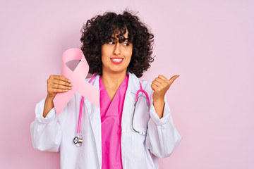 Young arab doctor woman with curly hair holding cancer ribbon over isolated pink background pointing and showing with thumb up to the side with happy face smiling