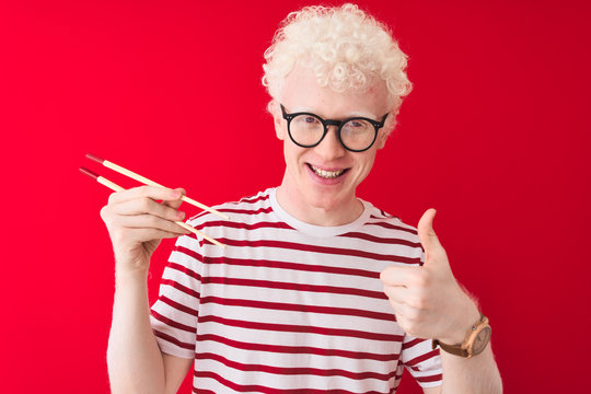 Young albino blond man holding chopsticks standing over isolated white background happy with big smile doing ok sign, thumb up with fingers, excellent sign