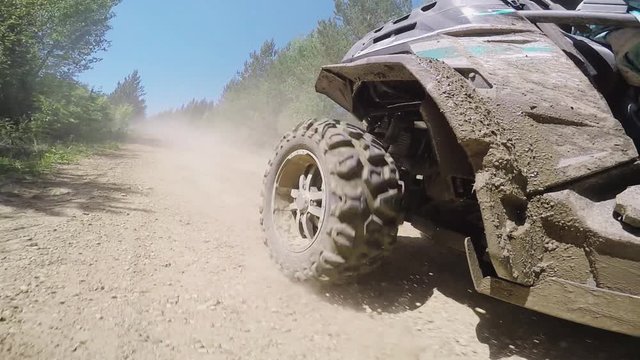 Side view of dirty extreme quadrocycle actively riding and moving along offroad among grasses, trees and bushes outside on sunny warm day.