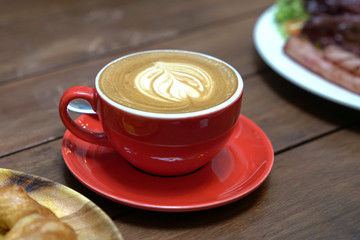 Flat white coffee with heart pattern in a red cup and par tial view of croissant and salad bacon.