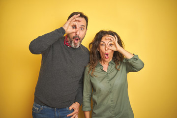 Beautiful middle age couple over isolated yellow background doing ok gesture shocked with surprised face, eye looking through fingers. Unbelieving expression.