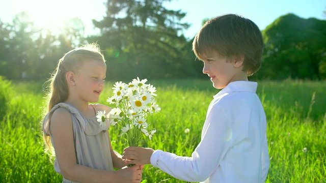 ittle cute boy giving to beautiful girl bouquet flowers chamomiles outdoors at sunset. first love, friendship, childhood, Funny kids, child. lovely childrens couple in nature, summer, slow-motion