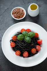 Squid-ink spaghetti with sun dried tomato pesto, fresh cherry tomatoes and green basil served in a white plate, vertical shot