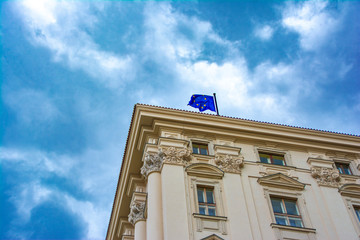 Part of the facade of the classic building of a public institution with the flag of the European Union on the roof against a blue sky.