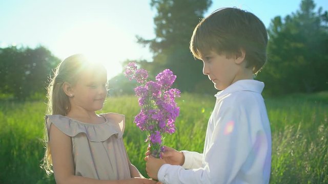 ittle cute boy giving to beautiful girl bouquet flowers peonies outdoors at sunset. first love, friendship, childhood, Funny kids, child. lovely childrens couple in nature, summer, slow-motion
