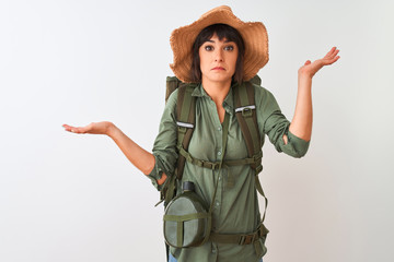 Hiker woman wearing backpack hat and water canteen over isolated white background clueless and confused expression with arms and hands raised. Doubt concept.