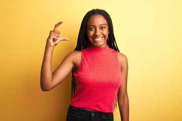 African american woman wearing red casual t-shirt standing over isolated yellow background smiling and confident gesturing with hand doing small size sign with fingers looking and the camera