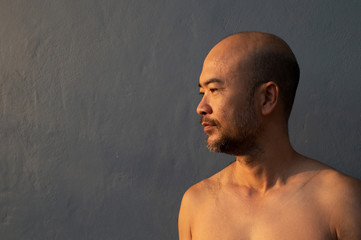 adult bald beard 40s Japanese man topless profile portrait with dark grey blue concrete wall...