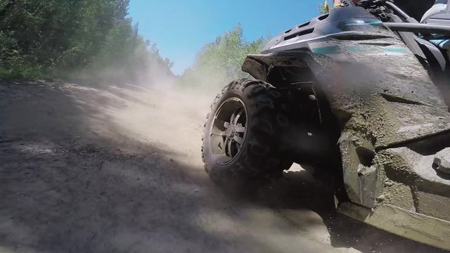 Quadrocycle is extremely riding and moving along dirty dusty offroad among grasses, trees and bushes outside on sunny warm day.