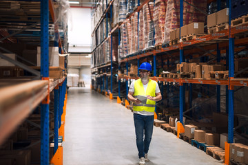 Warehouse worker looking at shelves with packages and walking through large warehouse storage distribution area.