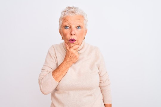 Senior grey-haired woman wearing turtleneck sweater standing over isolated white background Looking fascinated with disbelief, surprise and amazed expression with hands on chin