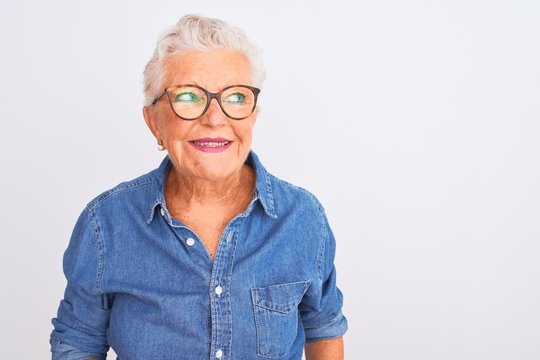 Senior grey-haired woman wearing denim shirt and glasses over isolated white background smiling looking to the side and staring away thinking.