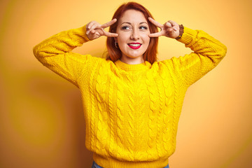 Beautiful redhead woman wearing winter sweater standing over isolated yellow background Doing peace symbol with fingers over face, smiling cheerful showing victory