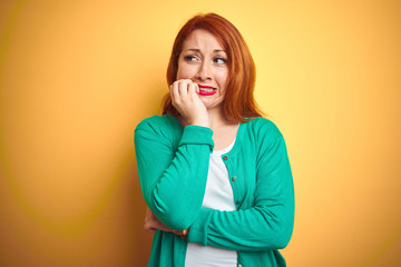 Youg beautiful redhead woman wearing winter green sweater over isolated yellow background looking stressed and nervous with hands on mouth biting nails. Anxiety problem.