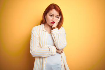 Beautiful redhead woman wearing striped shirt standing over isolated yellow background thinking looking tired and bored with depression problems with crossed arms.