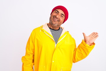 Middle age man wearing rain coat and woolen hat standing over isolated white background smiling cheerful presenting and pointing with palm of hand looking at the camera.