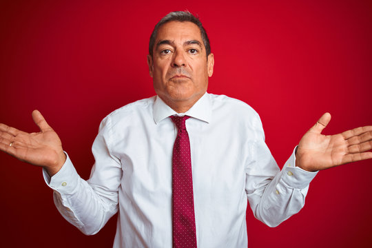Handsome middle age businessman standing over isolated red background clueless and confused expression with arms and hands raised. Doubt concept.