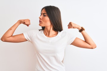 Young beautiful woman wearing casual t-shirt standing over isolated white background showing arms muscles smiling proud. Fitness concept.