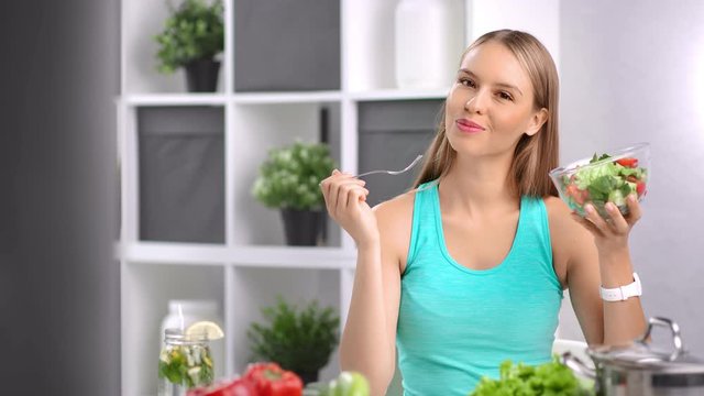 Attractive dieting girl eating fresh organic vegetable salad in kitchen at home looking at camera