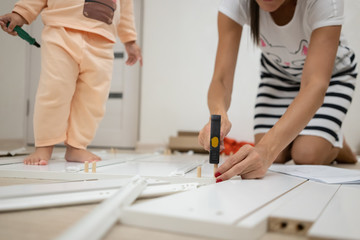 Mom and daughter collects a box. A woman is assembling a white wooden cabinet using a screwdriver.