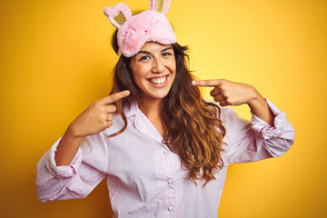 Young woman wearing pajama and sleep mask standing over yellow isolated background smiling cheerful showing and pointing with fingers teeth and mouth. Dental health concept.