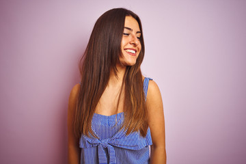 Young beautiful woman wearing striped dress standing over isolated pink background looking away to side with smile on face, natural expression. Laughing confident.