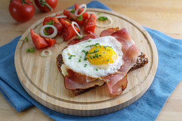 fried egg and ham on a dark whole meal bread, tomatoes, onion rings and herbs garnish, served on a kitchen board and a blue napkin