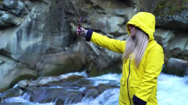 Young attractive blonde girl in yellow raincoat making selfie at the waterfall in mountains nature ecology student youth freedom lifestyle millenial model forest park autumn vibes background tourist