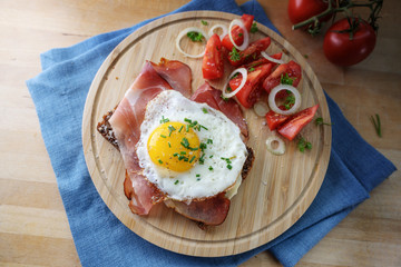 fried egg and ham on a dark whole meal bread, tomatoes, onion rings and herbs garnish, served on a kitchen board and a blue napkin, top view from above