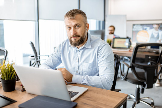 Portrait of a senior man dressed casually working on the laptop in the office with coworkers on the background