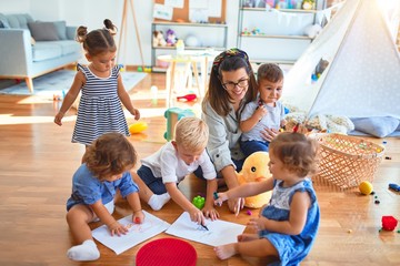 Beautiful teacher and group of toddlers sitting on the floor drawing using paper and pencil around lots of toys at kindergarten - obrazy, fototapety, plakaty