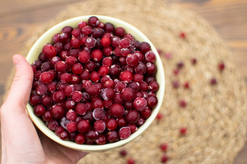 Female hand giving big bowl full of frozen cranberry at blurred wooden background with eco napkin, scattered berries