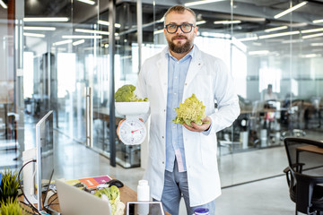Portrait of senior nutritionist in medical gown standing with green vegetables and weight in office