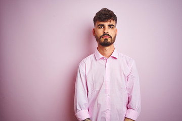 Obraz premium Young man with tattoo wearing shirt standing over isolated pink background with serious expression on face. Simple and natural looking at the camera.