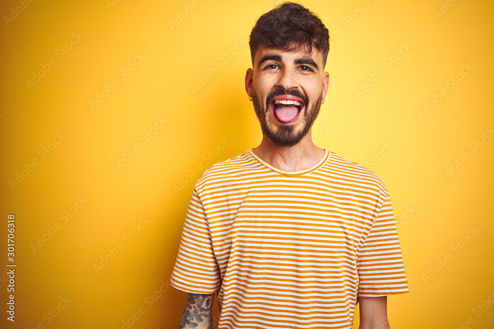 Poster young man with tattoo wearing striped t-shirt standing over isolated yellow background sticking tong