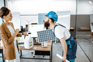 Male worker and woman engineer working on a project of alternative energy, holding wind turbine and solar panel model in the office