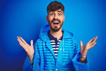 Young handsome man wearing rain coat standing over isolated blue background celebrating crazy and amazed for success with arms raised and open eyes screaming excited. Winner concept