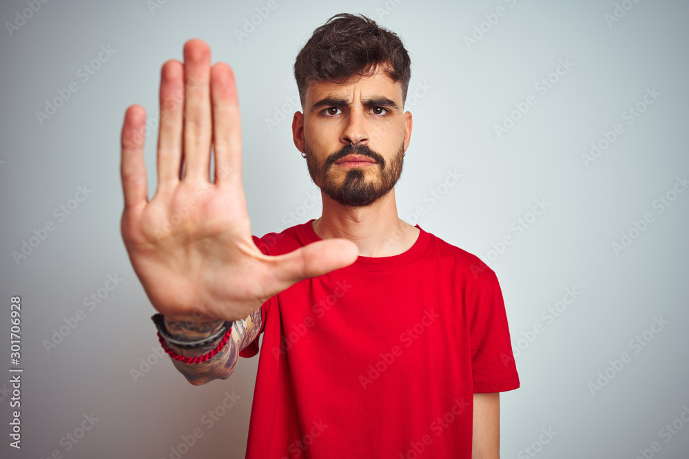 Sticker Young man with tattoo wearing red t-shirt standing over isolated white background doing stop sing with palm of the hand. Warning expression with negative and serious gesture on the face.