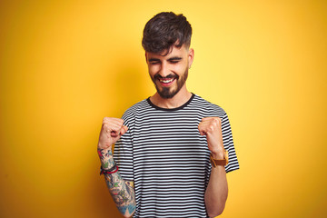 Young man with tattoo wearing striped t-shirt standing over isolated yellow background very happy and excited doing winner gesture with arms raised, smiling and screaming for success