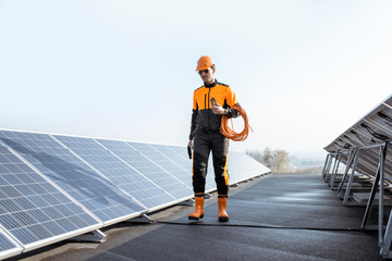 Well-equipped worker in protective orange clothing walking and examining solar panels on a photovoltaic rooftop plant. Concept of maintenance and installation of solar stations