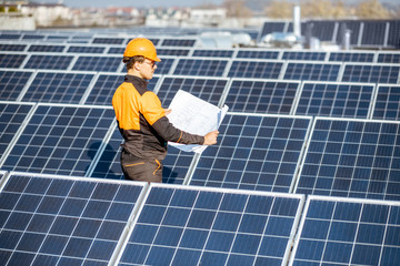 Engineer in protective workwear with blueprints of solar station project on a photovoltaic rooftop plant. Concept of designing power plants