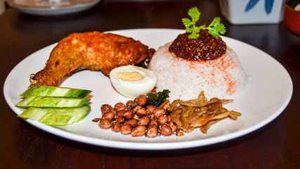 Malaysian Food Concept : traditional food of Malaysia called "Nasi Lemak" served rice with fried chicken, fish, nuts, Chilli paste, egg and slice cucumber in white plate on wood table in restaurant