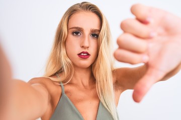 Obraz premium Young beautiful woman wearing t-shirt make selfie by camera over isolated white background with angry face, negative sign showing dislike with thumbs down, rejection concept