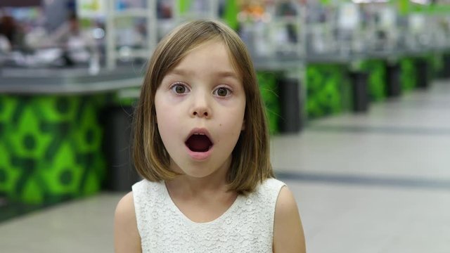 Close-up portrait of a girl with blond hair, 5-6 years old. Different emotions on the face of the model. A child is standing in a shopping center or supermarket against the background.