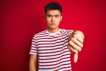 Young asian chinese man wearing striped t-shirt standing over isolated red background looking unhappy and angry showing rejection and negative with thumbs down gesture. Bad expression.