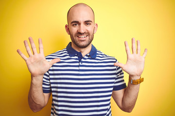 Young bald man with beard wearing casual striped blue t-shirt over yellow isolated background showing and pointing up with fingers number ten while smiling confident and happy.