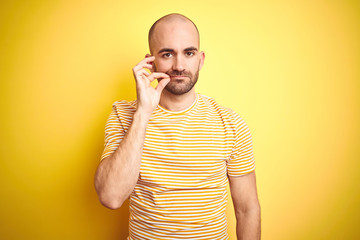 Young bald man with beard wearing casual striped t-shirt over yellow isolated background mouth and...