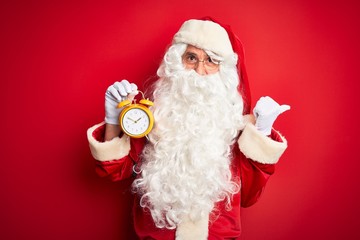 Senior man wearing Santa Claus costume holding alarm clock over isolated red background pointing and showing with thumb up to the side with happy face smiling