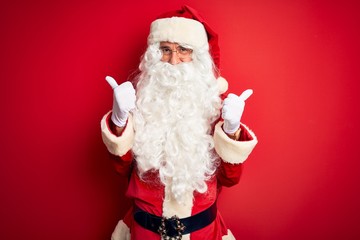 Middle age handsome man wearing Santa costume standing over isolated red background success sign doing positive gesture with hand, thumbs up smiling and happy. Cheerful expression and winner gesture.