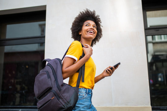 happy young black woman walking outside with cellphone and bag